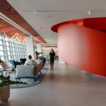 Modern open-plan office lounge with a large red curved architectural feature, seating area, and floor-to-ceiling windows.