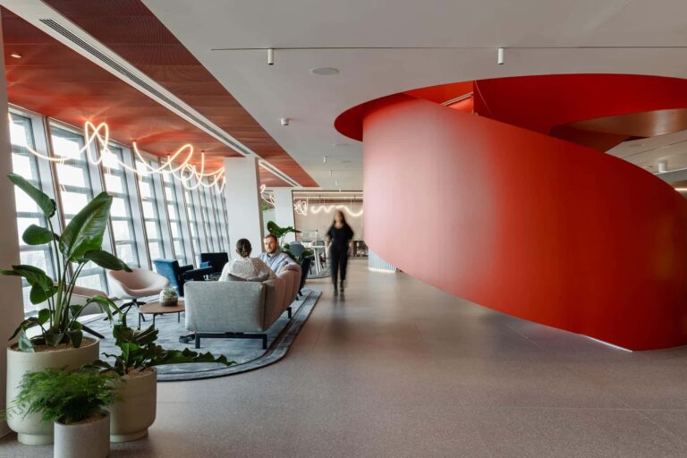 Modern open-plan office lounge with a large red curved architectural feature, seating area, and floor-to-ceiling windows.