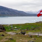 A Greenlandic flag flying over a coastal settlement with icebergs, a fjord, and snow-capped mountains in the background.