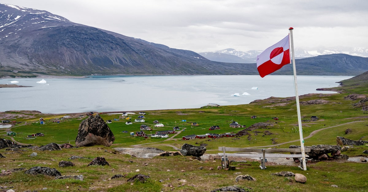 A Greenlandic flag flying over a coastal settlement with icebergs, a fjord, and snow-capped mountains in the background.