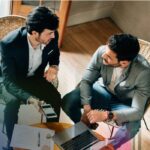 Two businessmen in a meeting discussing documents and a laptop at a small table