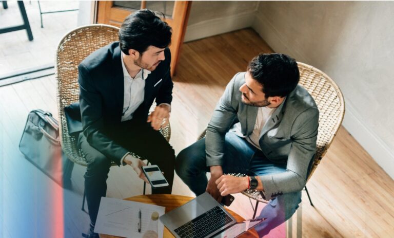 Two businessmen in a meeting discussing documents and a laptop at a small table