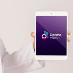 A hand holding a tablet displaying the Optima Health company logo on a purple screen