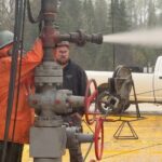 Workers in protective gear monitor a wellhead during a pressure/flow test with gas venting from the pipe.