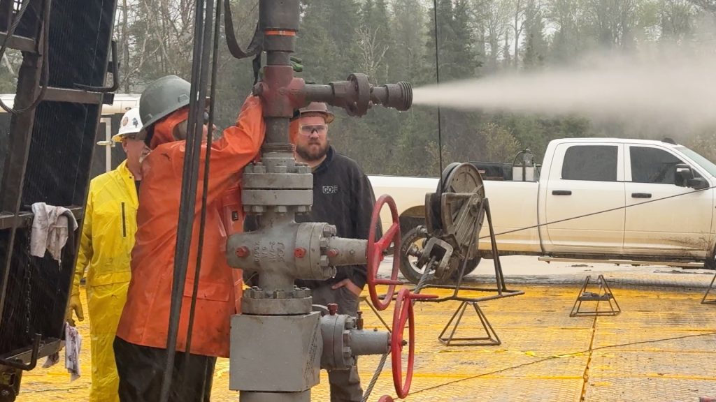 Workers in protective gear monitor a wellhead during a pressure/flow test with gas venting from the pipe.