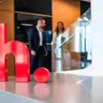Large red 'h.' sculpture on a reception desk at Hikma Pharmaceuticals' MENA headquarters, with colleagues talking in the background.