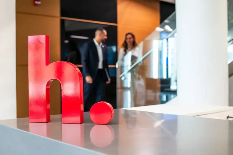 Large red 'h.' sculpture on a reception desk at Hikma Pharmaceuticals' MENA headquarters, with colleagues talking in the background.