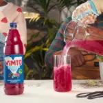 A bottle of Vimto no-added-sugar squash on a table beside a glass being poured with the drink, in a garden-themed lifestyle scene.