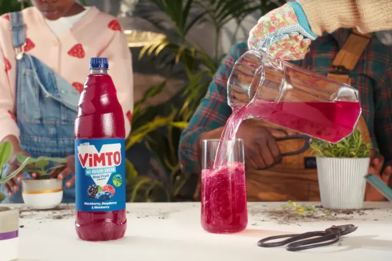 A bottle of Vimto no-added-sugar squash on a table beside a glass being poured with the drink, in a garden-themed lifestyle scene.