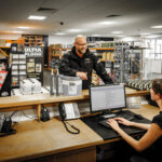 Staff at a Headlam Group trade counter serving a customer, surrounded by flooring adhesive products and warehouse stock.