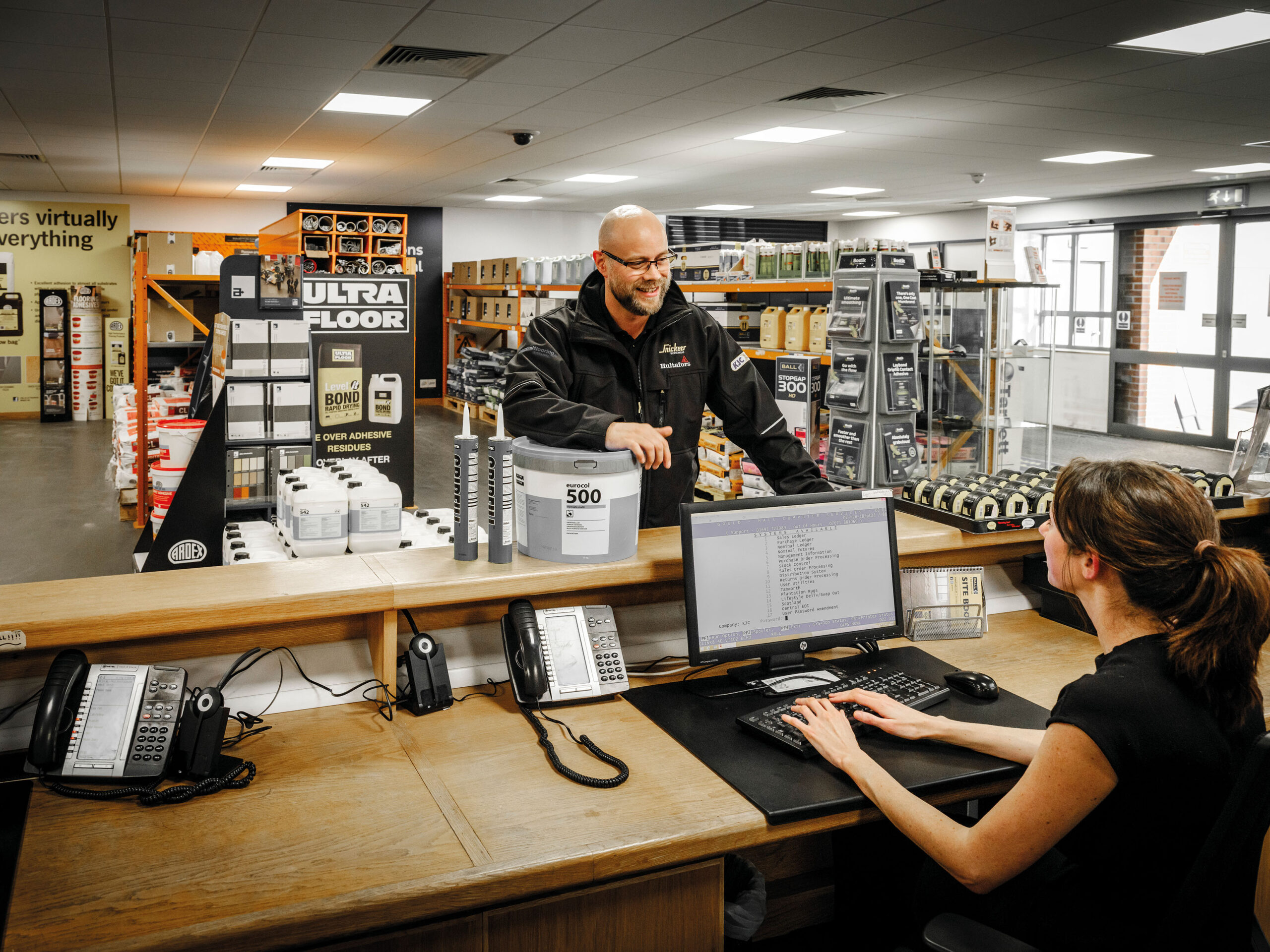 Staff at a Headlam Group trade counter serving a customer, surrounded by flooring adhesive products and warehouse stock.