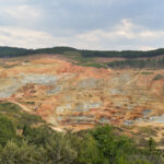 Aerial view of a large open-pit copper and gold mine with terraced rock benches surrounded by forested hills.