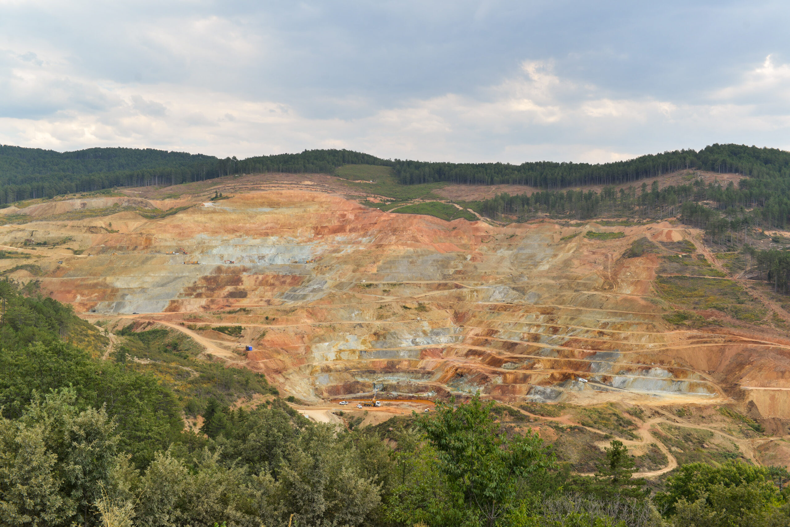 Aerial view of a large open-pit copper and gold mine with terraced rock benches surrounded by forested hills.