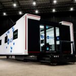 A white production office trailer branded with the 'facilities by ADF' logo, parked inside a large industrial hangar with exposed steel roof trusses and overhead lighting.