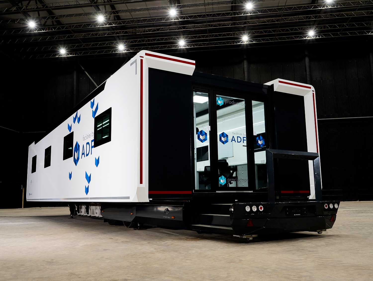 A white production office trailer branded with the 'facilities by ADF' logo, parked inside a large industrial hangar with exposed steel roof trusses and overhead lighting.