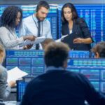 Financial analysts reviewing documents in a trading room surrounded by screens showing market data and charts