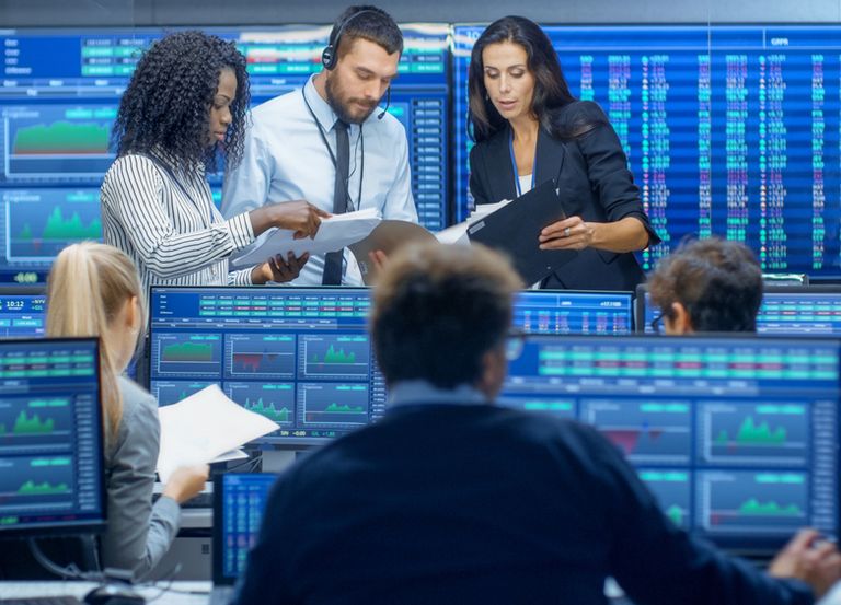 Financial analysts reviewing documents in a trading room surrounded by screens showing market data and charts