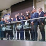 Group of business people applauding on a balcony overlooking the London Stock Exchange trading floor, with 'LONDON STOCK EXCHANGE' signage visible on the glass railing.