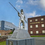 Stainless-steel steelworker statue in front of a red-brick building with a 'Goodwin PLC' sign