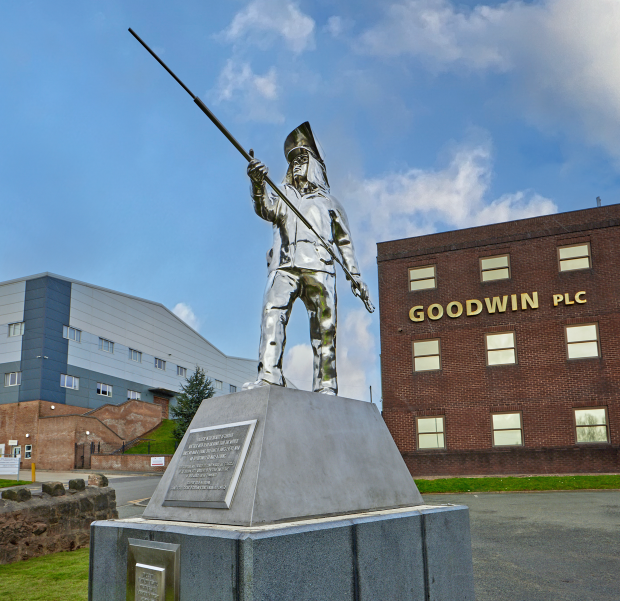 Stainless-steel steelworker statue in front of a red-brick building with a 'Goodwin PLC' sign
