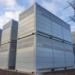Rows of Invinity Energy Systems vanadium flow battery storage units installed at a grid-connected energy storage site, with electricity transmission pylons in the background.