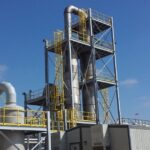 Industrial chemical processing plant with tall metal extraction towers and yellow safety railings under a blue sky.
