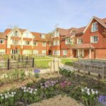 Landscaped garden courtyard with lawns, flowerbeds and benches in front of a red-brick residential retirement/senior living building.