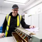 Smiling factory worker in a high-visibility vest and safety cap operating printing/finishing machinery