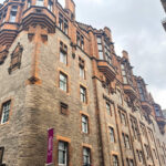 Exterior view of a stone and sandstone building with a vertical maroon 'Safestay hostels & hotels' sign, looking up from street level toward a partly cloudy sky.