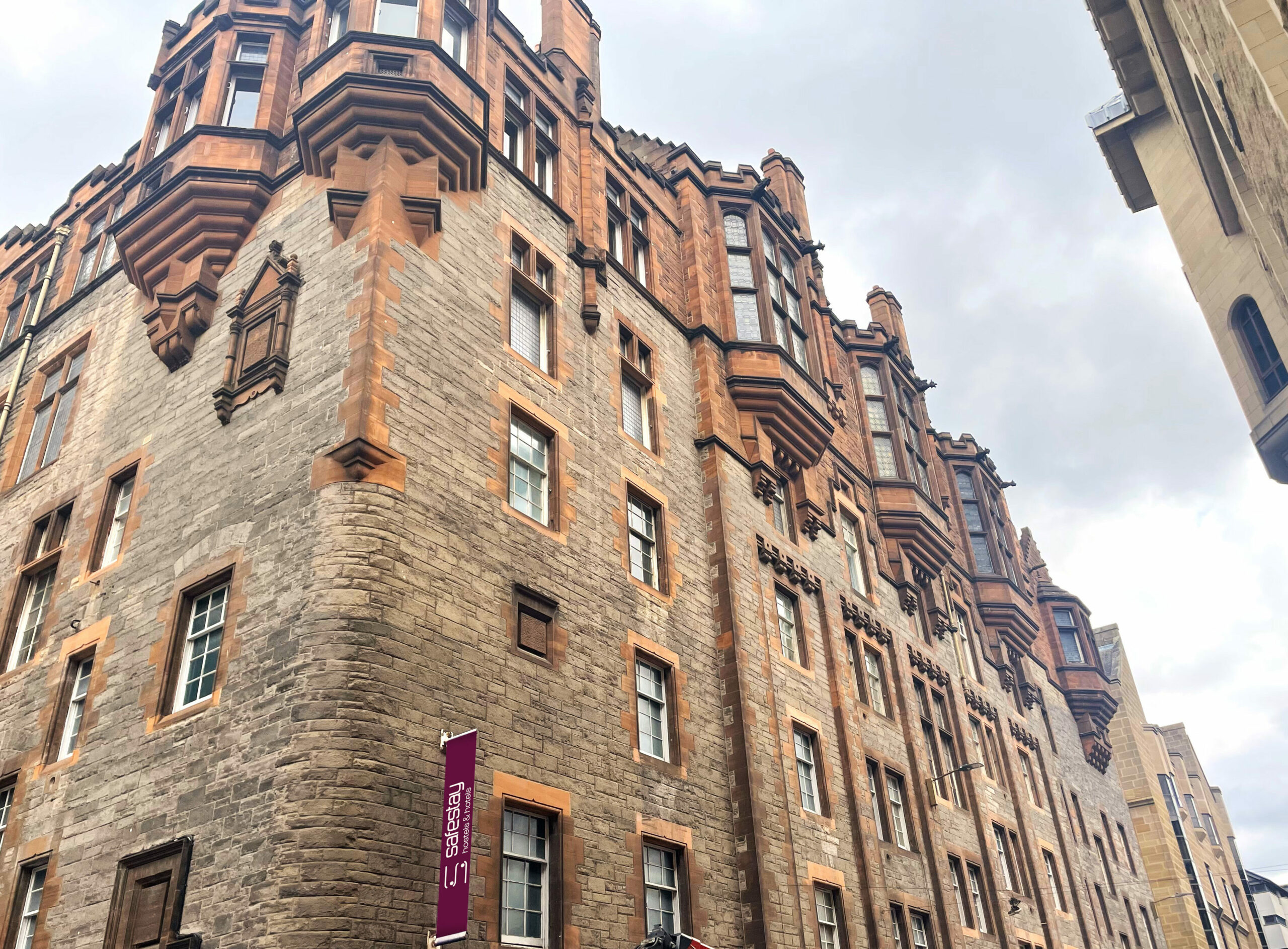 Exterior view of a stone and sandstone building with a vertical maroon 'Safestay hostels & hotels' sign, looking up from street level toward a partly cloudy sky.