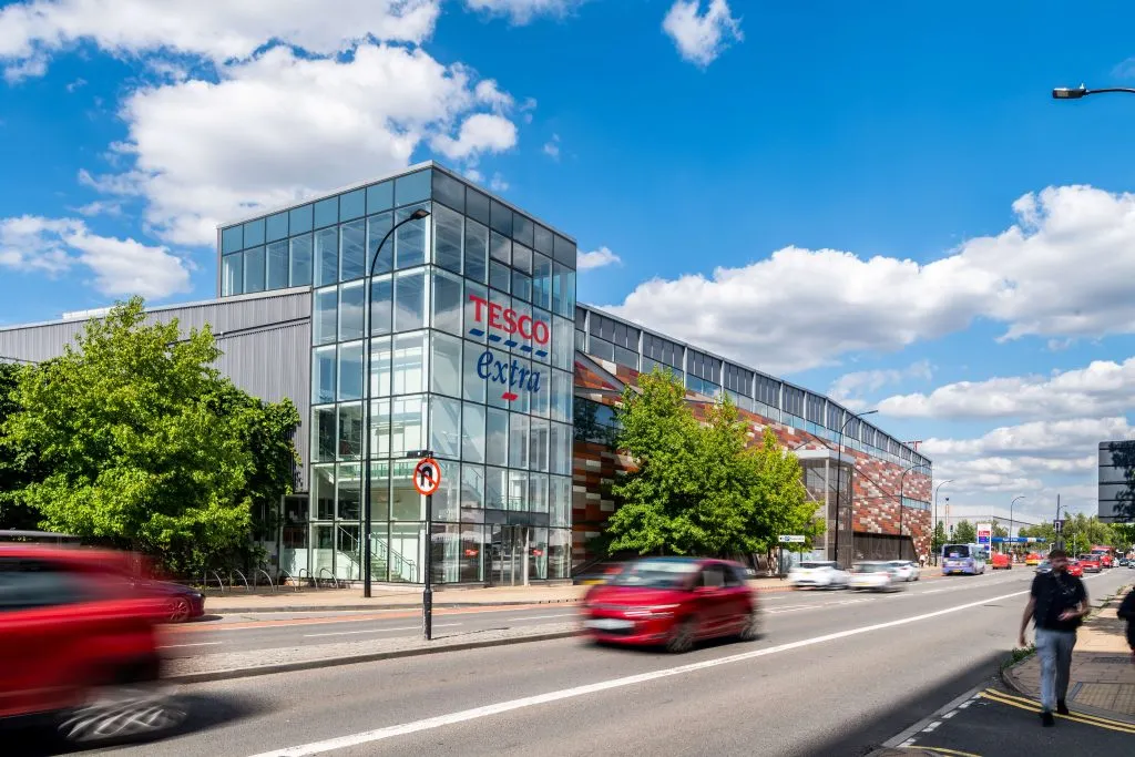 Exterior view of a large Tesco Extra supermarket building with a busy road in the foreground, representative of the type of retail property held by Supermarket Income REIT.