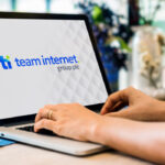 Close-up of a laptop screen displaying the 'team internet. group plc' logo while a person's hands type on the keyboard, with a blurred indoor background