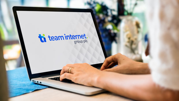 Close-up of a laptop screen displaying the 'team internet. group plc' logo while a person's hands type on the keyboard, with a blurred indoor background