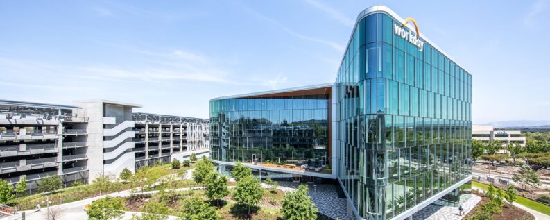 Aerial view of a modern glass office building with the Workday logo on its roofline, beside a landscaped courtyard and parking structure