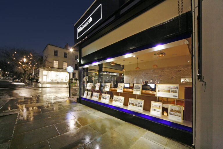 Exterior night view of a Winkworth estate agency branch with illuminated sign and window displaying property listings.