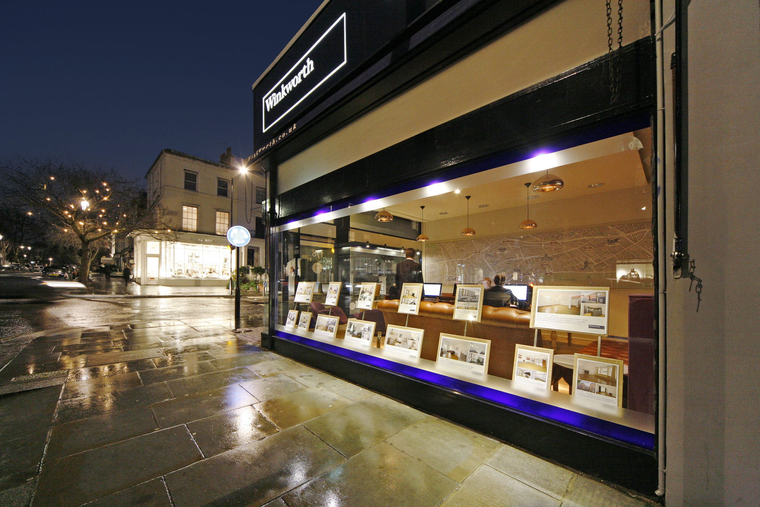 Exterior night view of a Winkworth estate agency branch with illuminated sign and window displaying property listings.