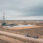 Aerial view of an oil drilling rig and associated equipment in a flat, arid Permian Basin landscape, with a flatbed truck driving on a dirt road in the foreground and a second rig visible in the distance under an overcast sky