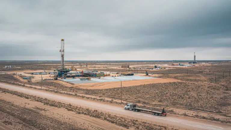 Aerial view of an oil drilling rig and associated equipment in a flat, arid Permian Basin landscape, with a flatbed truck driving on a dirt road in the foreground and a second rig visible in the distance under an overcast sky