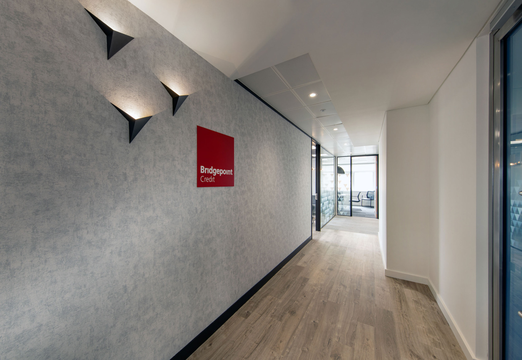 Modern office corridor with wood flooring and a red 'Bridgepoint Credit' wall sign, glass-walled meeting rooms visible in the background.