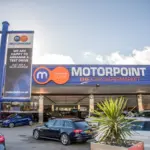 Exterior of a Motorpoint car supermarket showroom with branded signage, a pole sign reading 'Motorpoint - The Car Supermarket', and rows of used cars parked on the forecourt under a partly cloudy sky.