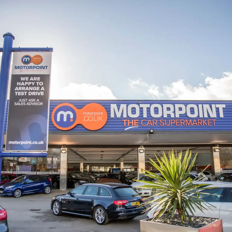 Exterior of a Motorpoint car supermarket showroom with branded signage, a pole sign reading 'Motorpoint - The Car Supermarket', and rows of used cars parked on the forecourt under a partly cloudy sky.