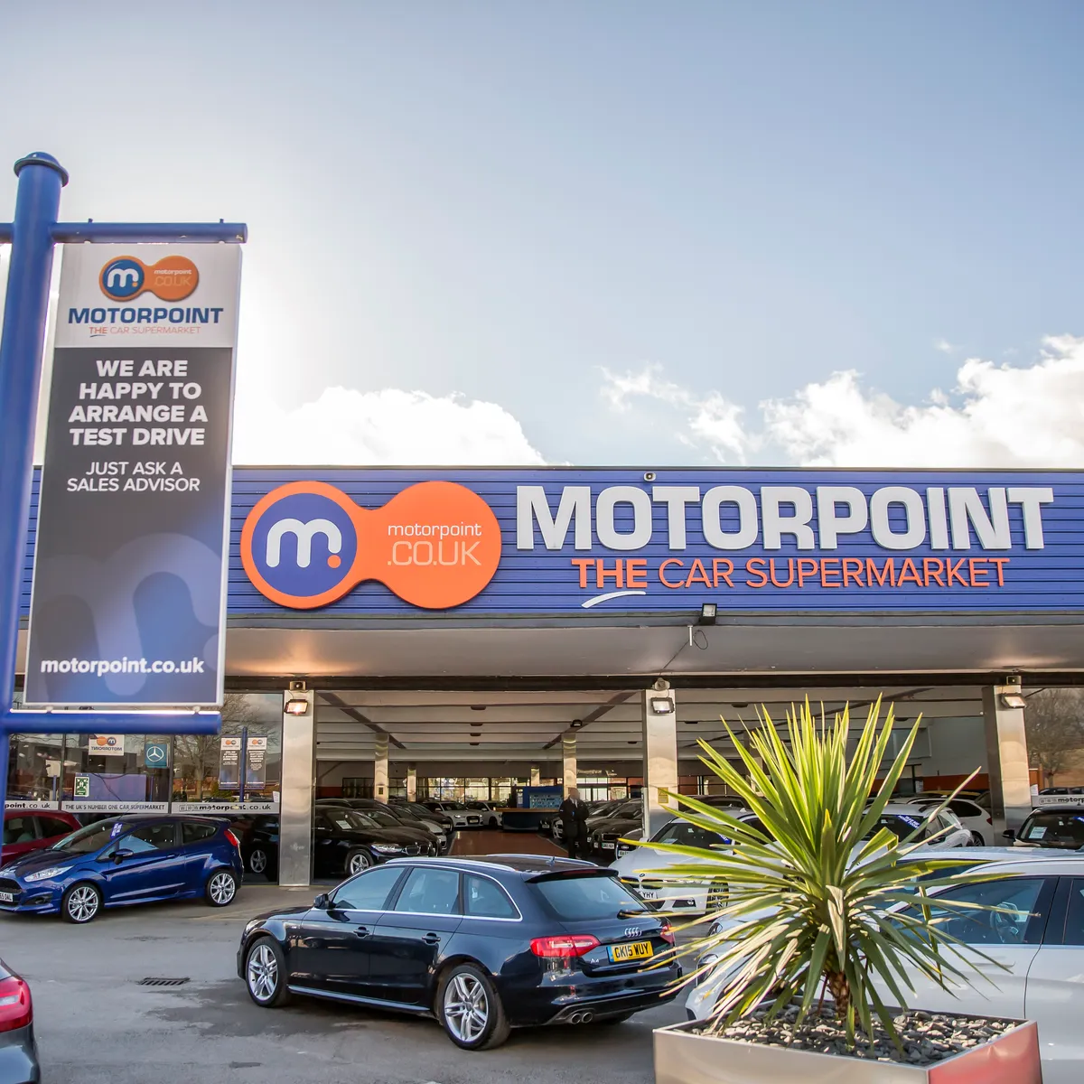 Exterior of a Motorpoint car supermarket showroom with branded signage, a pole sign reading 'Motorpoint - The Car Supermarket', and rows of used cars parked on the forecourt under a partly cloudy sky.