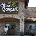 Exterior of an Olive Garden Italian Kitchen restaurant with stone facade, showing the branded sign, entrance doors, a customer walking in, and an accessible parking sign.