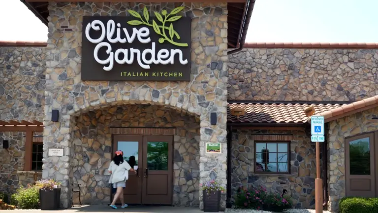 Exterior of an Olive Garden Italian Kitchen restaurant with stone facade, showing the branded sign, entrance doors, a customer walking in, and an accessible parking sign.