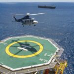 Helicopter landing on an offshore oil platform helideck with a support vessel visible on the horizon.