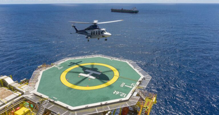 Helicopter landing on an offshore oil platform helideck with a support vessel visible on the horizon.