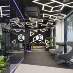 Interior of Entain's London office lobby featuring an illuminated purple 'Entain' sign, repeated Entain sigma-symbol motifs on glass panels, geometric ceiling lighting, potted plants, and grey lounge chairs.