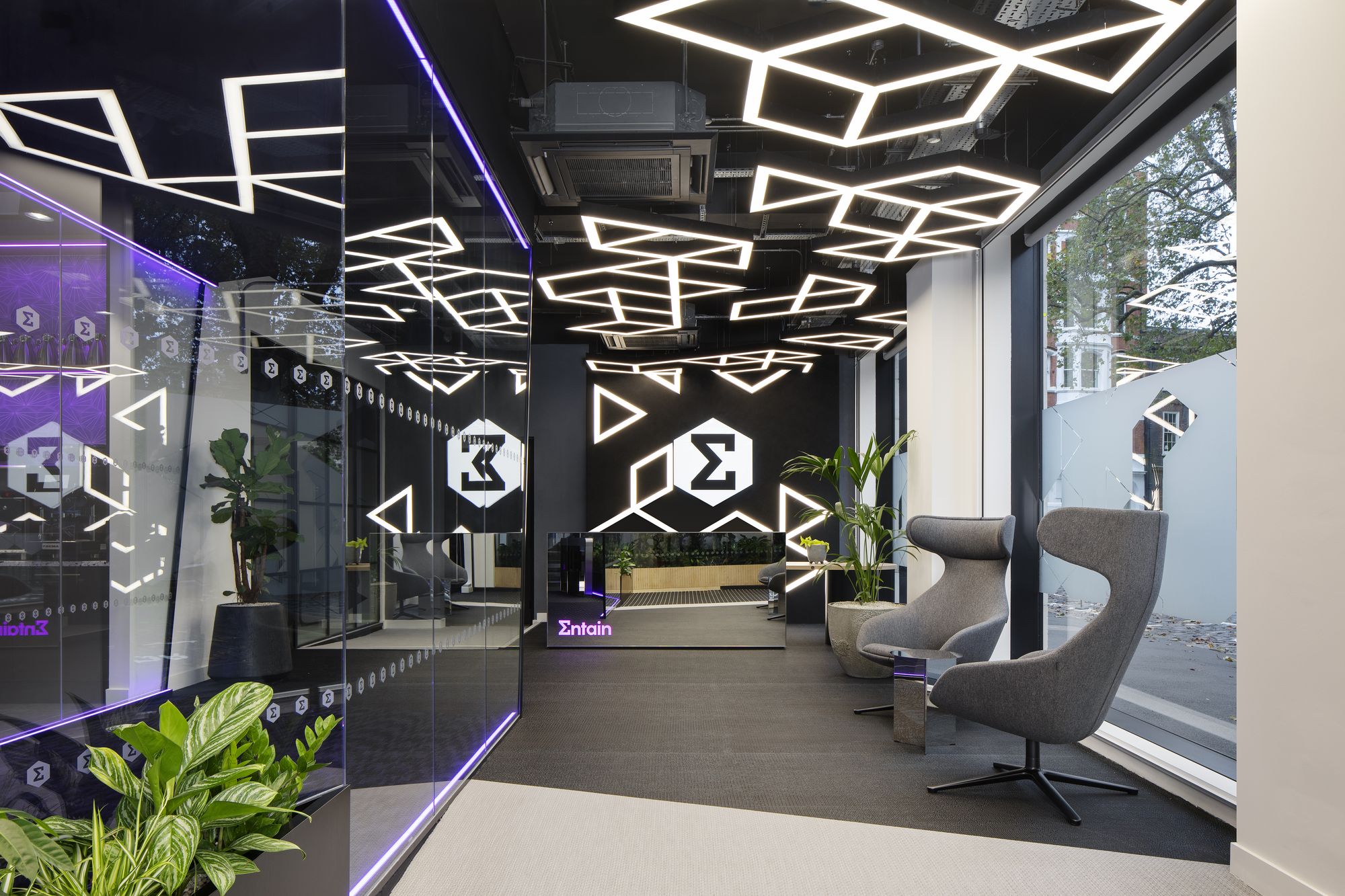 Interior of Entain's London office lobby featuring an illuminated purple 'Entain' sign, repeated Entain sigma-symbol motifs on glass panels, geometric ceiling lighting, potted plants, and grey lounge chairs.