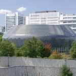 Exterior view of the Novartis Campus in Basel, showing the elliptical glass-mesh Novartis Pavillon building in front of office towers with the Novartis logo visible on a building facade, surrounded by trees and a stone retaining wall.