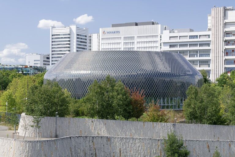 Exterior view of the Novartis Campus in Basel, showing the elliptical glass-mesh Novartis Pavillon building in front of office towers with the Novartis logo visible on a building facade, surrounded by trees and a stone retaining wall.